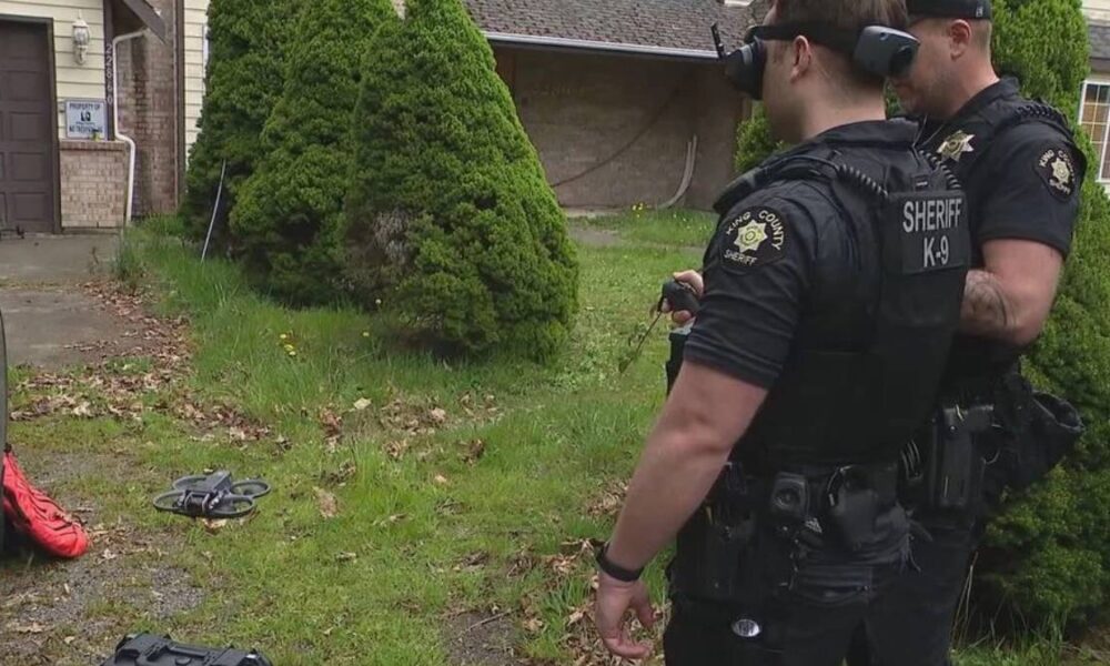Two sheriff officers in tactical gear stand in a residential yard, a small drone lies on the grass nearby.