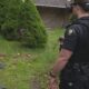 Two sheriff officers in tactical gear stand in a residential yard, a small drone lies on the grass nearby.