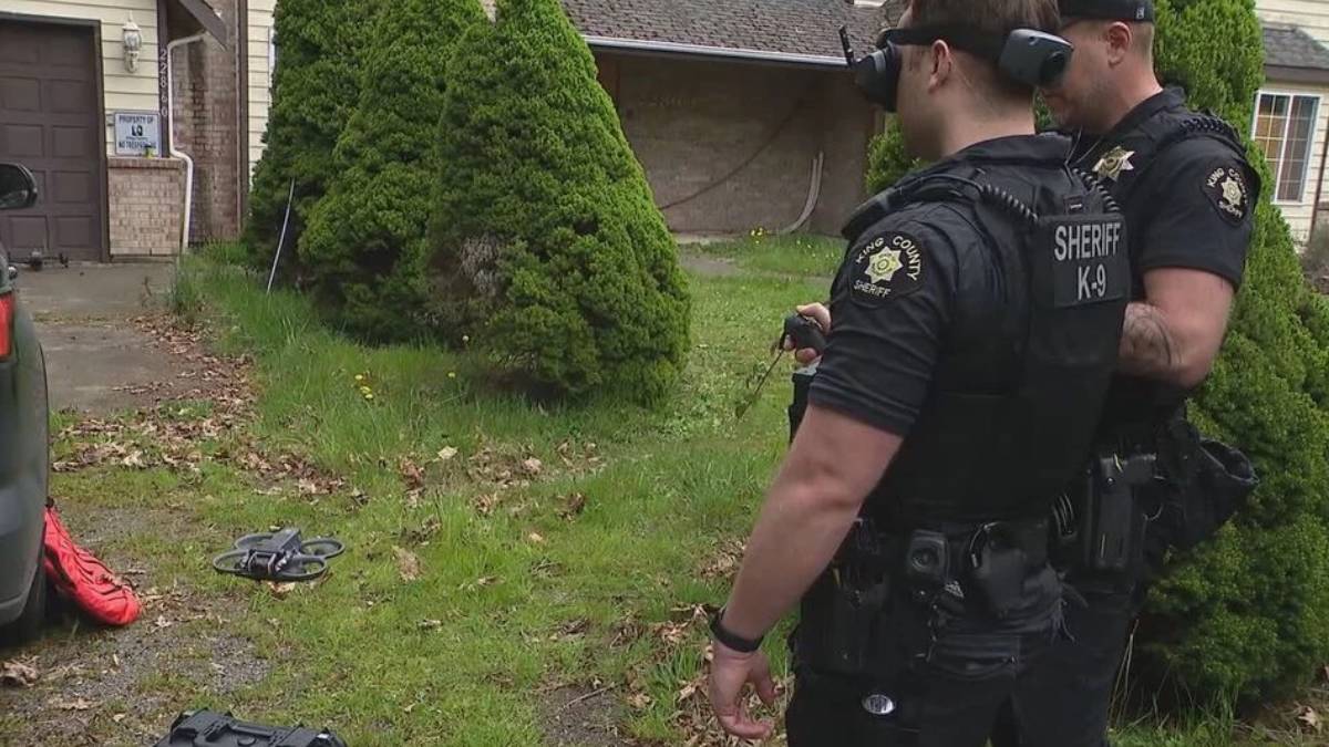 Two sheriff officers in tactical gear stand in a residential yard, a small drone lies on the grass nearby.