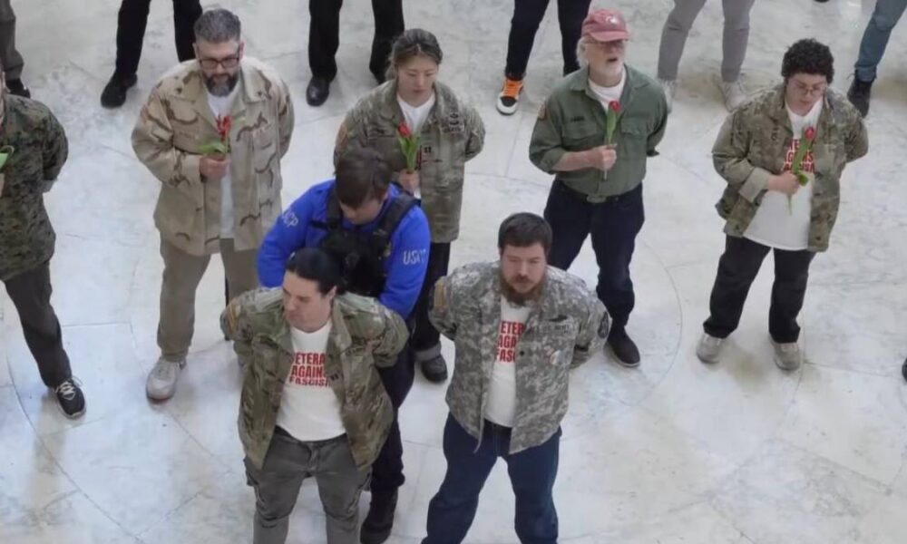 Group of veterans in camouflage jackets standing in a circle indoors, each holding a red rose.