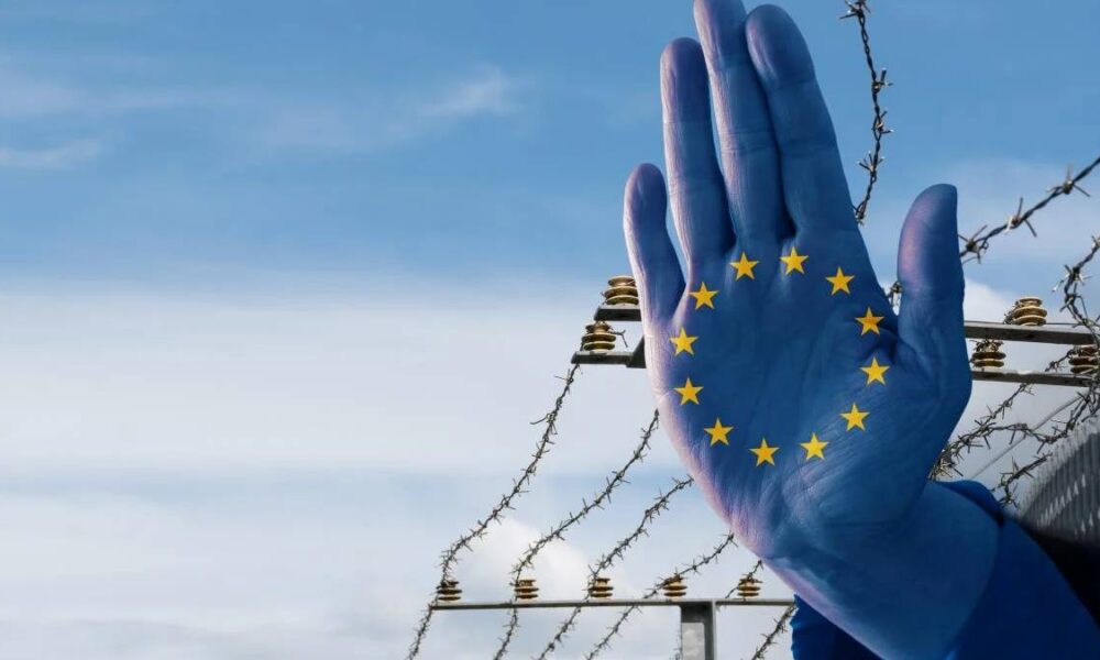 Blue glove with the European Union stars on the palm raised in front of a barbed‑wire border fence against a blue sky, symbolizing EU border issues.