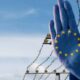Blue glove with the European Union stars on the palm raised in front of a barbed‑wire border fence against a blue sky, symbolizing EU border issues.