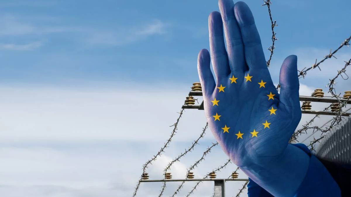 Blue glove with the European Union stars on the palm raised in front of a barbed‑wire border fence against a blue sky, symbolizing EU border issues.