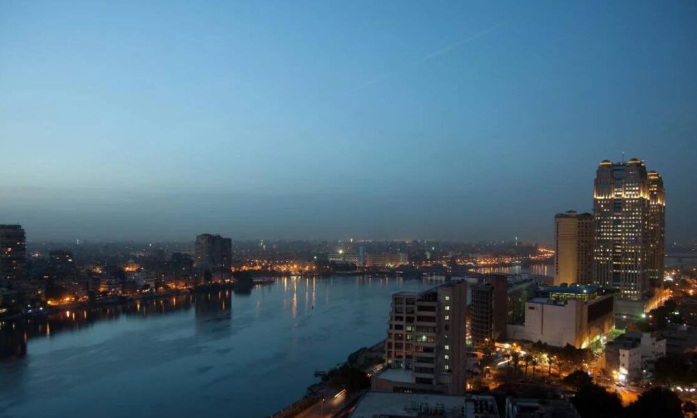 City skyline at dusk with a calm river reflecting orange and blue lights along the banks, tall illuminated buildings on the right.