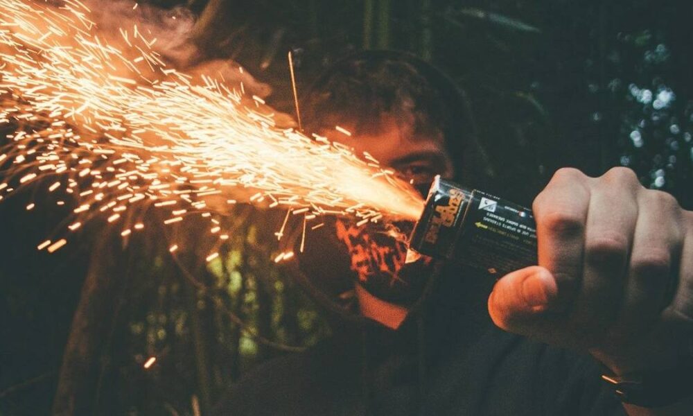 Person holding a handheld spark-emitting device, sparks shooting outward in a dark outdoor setting.