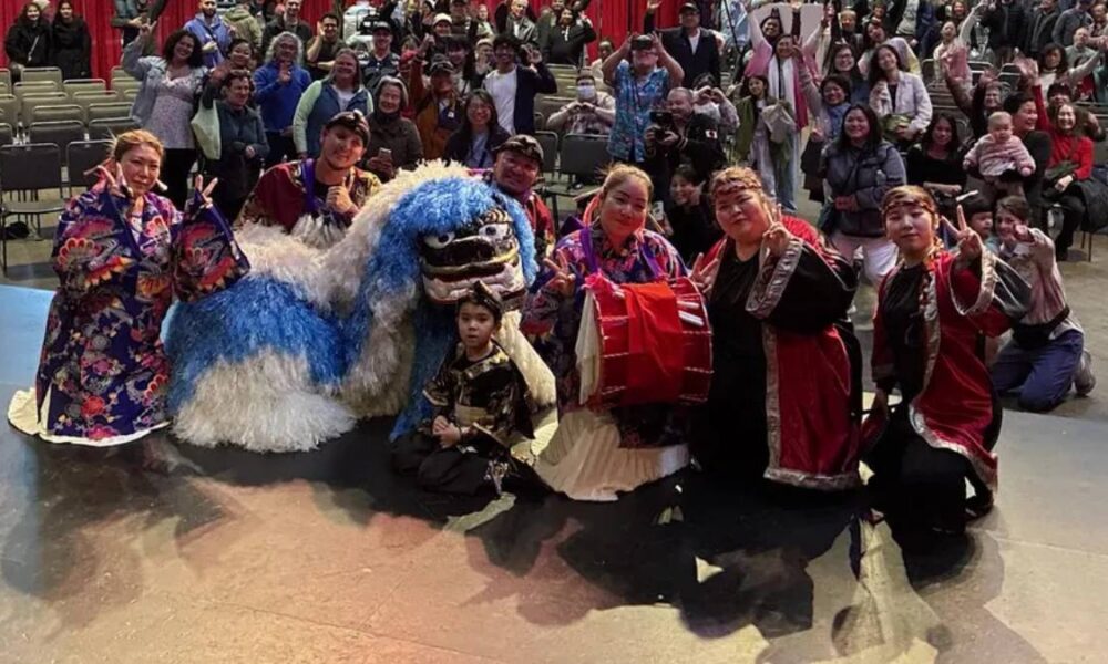 Group of performers posing with a blue-and-white dragon puppet on stage, a child in front, audience in the background.