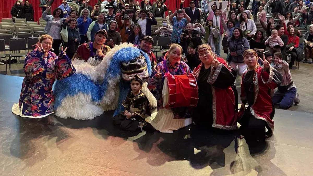 Group of performers posing with a blue-and-white dragon puppet on stage, a child in front, audience in the background.
