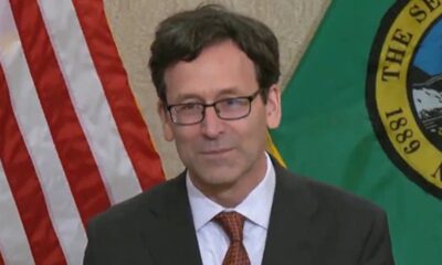 Man in a dark suit and red tie, wearing glasses, at a podium with the American flag and a green state flag in the background