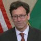 Man in a dark suit and red tie, wearing glasses, at a podium with the American flag and a green state flag in the background