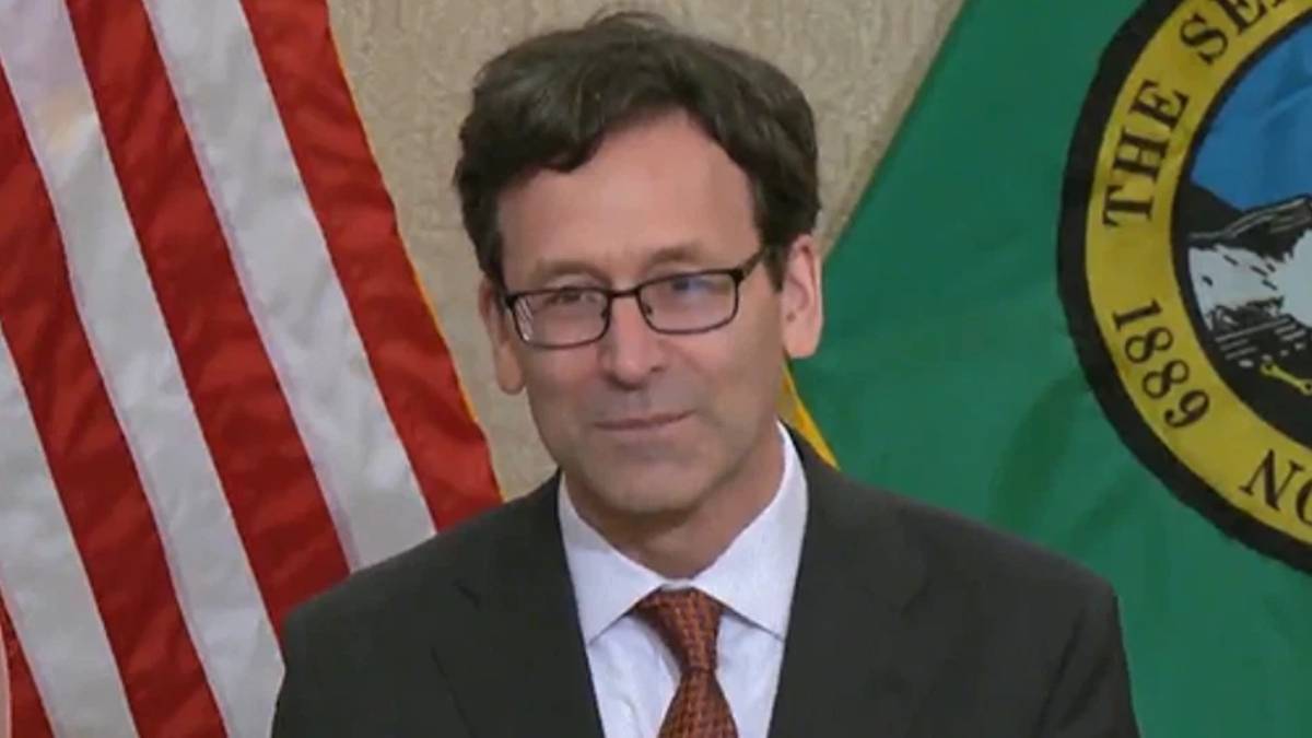 Man in a dark suit and red tie, wearing glasses, at a podium with the American flag and a green state flag in the background