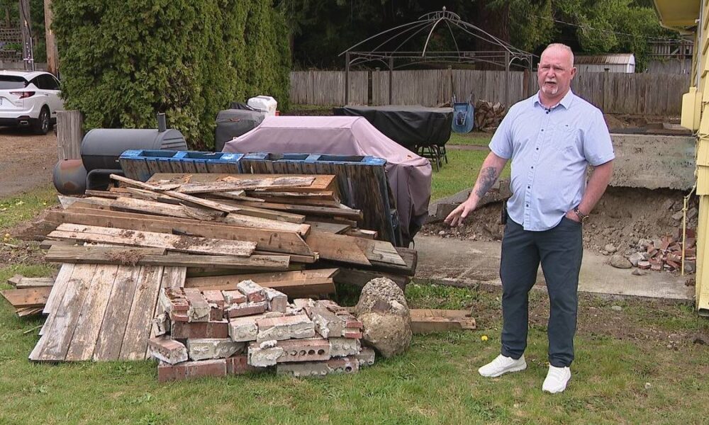 Man in a light shirt and dark pants stands in a backyard beside a large pile of weathered wooden planks and bricks, with a yellow house to the right.