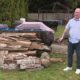 Man in a light shirt and dark pants stands in a backyard beside a large pile of weathered wooden planks and bricks, with a yellow house to the right.