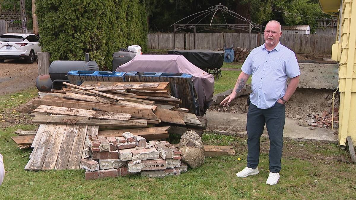 Man in a light shirt and dark pants stands in a backyard beside a large pile of weathered wooden planks and bricks, with a yellow house to the right.