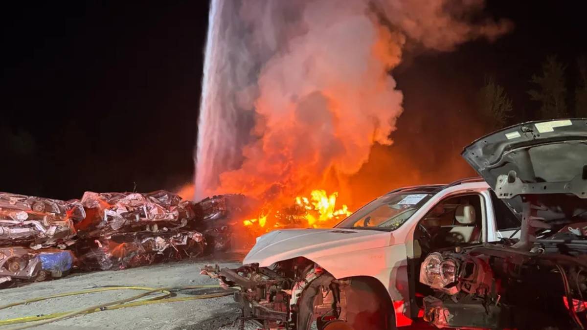 Nighttime junkyard scene with a burning pile of cars; orange flames and thick smoke rise behind a damaged white pickup truck.@] }? Wait remove extra.