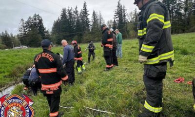 Firefighters in turnout gear line a grassy ditch for a water rescue drill, with ropes and equipment nearby.