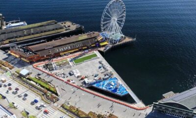 Aerial view of a waterfront pier with a large white Ferris wheel at the end of the dock and surrounding buildings along the water.
