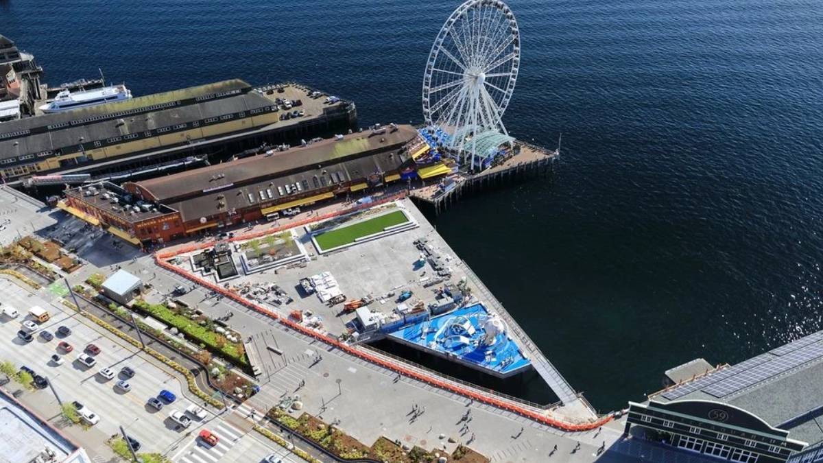 Aerial view of a waterfront pier with a large white Ferris wheel at the end of the dock and surrounding buildings along the water.