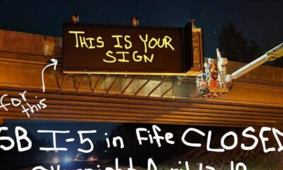 Nighttime highway work: workers in a bucket lift install a large sign above the freeway, with handwritten message 'THIS IS YOUR SIGN'. (Informative)