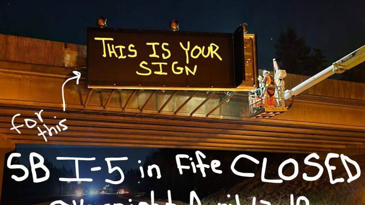 Nighttime highway work: workers in a bucket lift install a large sign above the freeway, with handwritten message 'THIS IS YOUR SIGN'. (Informative)