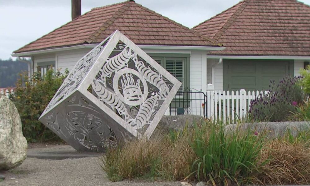 Decorative metal cube sculpture tilted on its corner in a residential yard, with intricate cut-out patterns against a white house and garden.