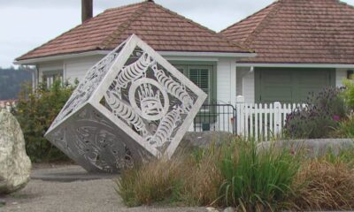 Decorative metal cube sculpture tilted on its corner in a residential yard, with intricate cut-out patterns against a white house and garden.