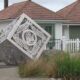 Decorative metal cube sculpture tilted on its corner in a residential yard, with intricate cut-out patterns against a white house and garden.