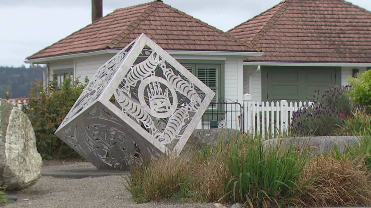 Decorative metal cube sculpture tilted on its corner in a residential yard, with intricate cut-out patterns against a white house and garden.