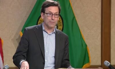 Man in a dark blazer and glasses speaking at a podium with flags in the background.