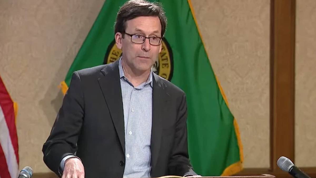 Man in a dark blazer and glasses speaking at a podium with flags in the background.