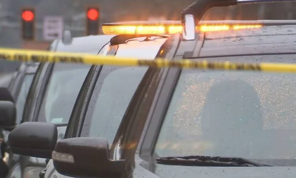Police cars with flashing orange lights at a scene, yellow caution tape across the foreground, rainy street in the background