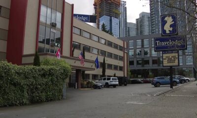 Low-rise hotel with Travelodge sign and multiple flags in front, parking lot and tall buildings in a city under construction.