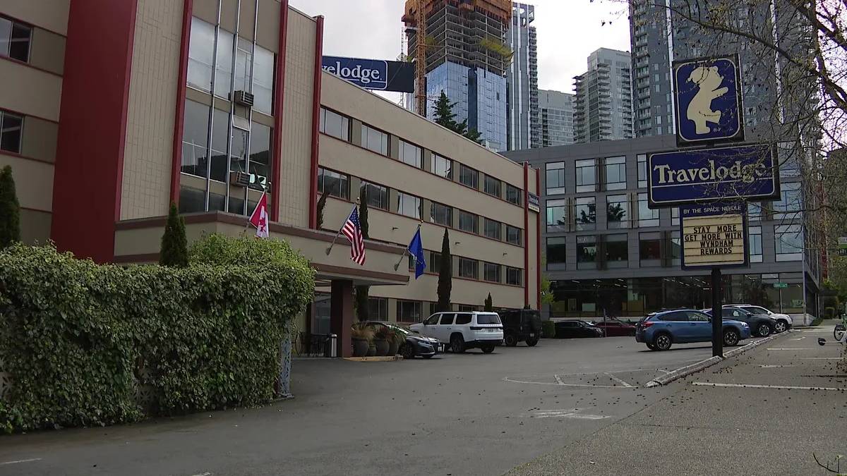 Low-rise hotel with Travelodge sign and multiple flags in front, parking lot and tall buildings in a city under construction.