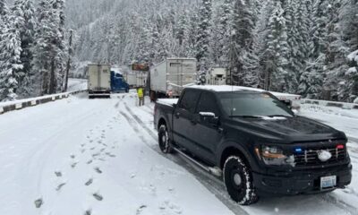 Black pickup truck with police lights on a snowy mountain road, where trucks are stalled and a worker in a high-visibility jacket stands nearby.
