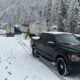 Black pickup truck with police lights on a snowy mountain road, where trucks are stalled and a worker in a high-visibility jacket stands nearby.