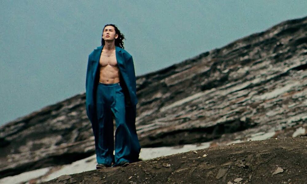 Man wearing a blue suit with the jacket open, no shirt, standing on a rocky hillside and looking upward at the sky.