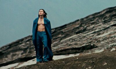 Man wearing a blue suit with the jacket open, no shirt, standing on a rocky hillside and looking upward at the sky.