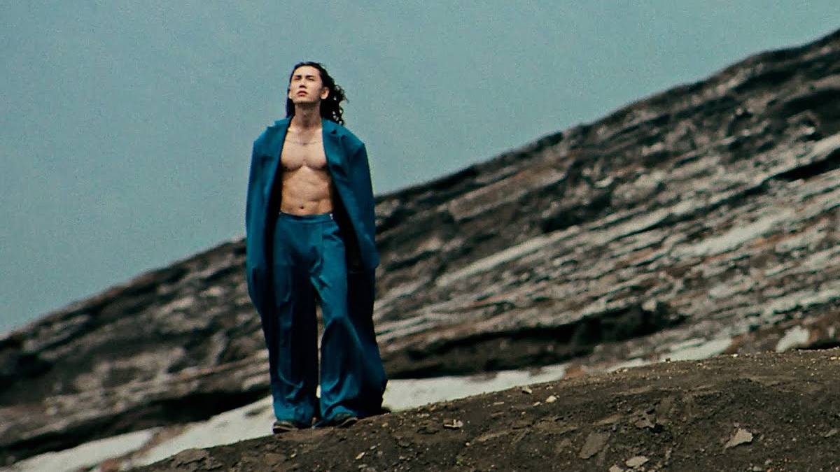 Man wearing a blue suit with the jacket open, no shirt, standing on a rocky hillside and looking upward at the sky.