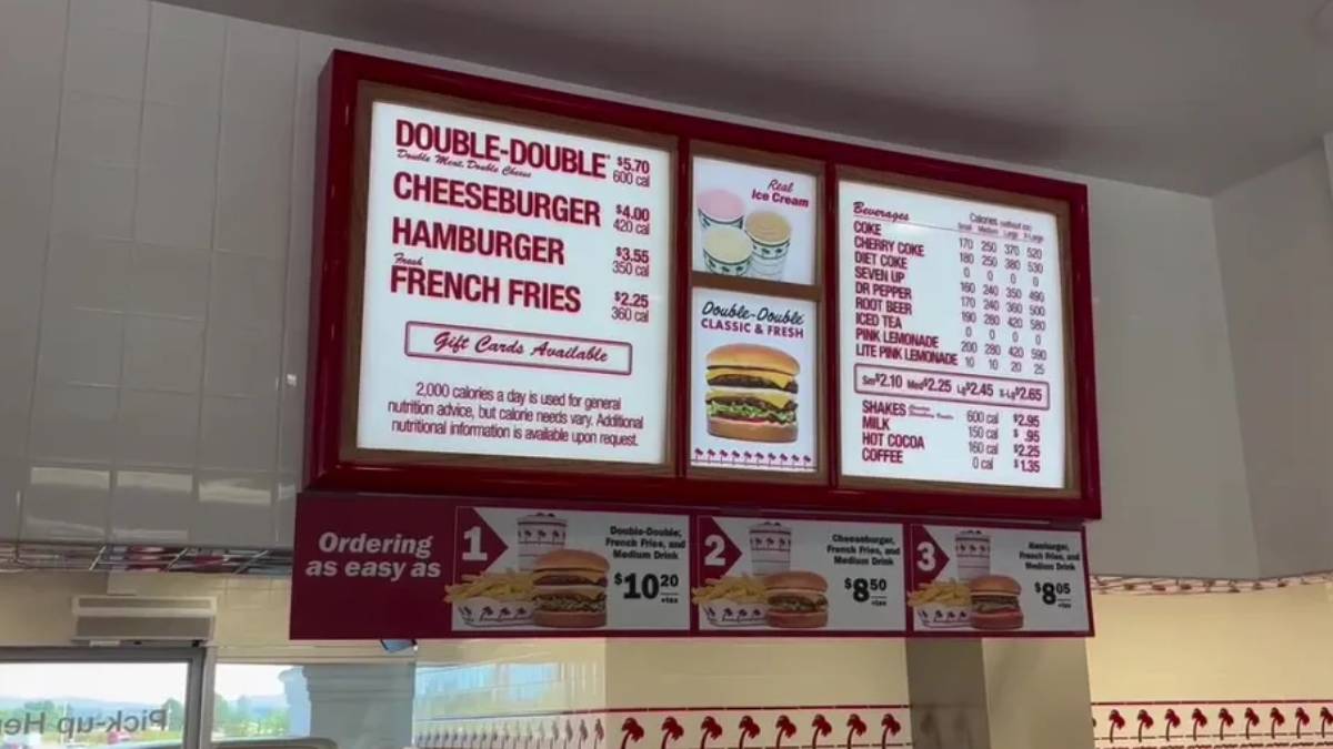 Illuminated fast-food menu board in a red frame listing Double-Double, Cheeseburger, Hamburger, French Fries with prices; below are ordering steps and burger images.