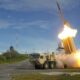 Military rocket launcher fires a missile, producing a bright flame and smoke on a test range.