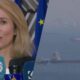 Woman speaking at a press conference with multiple microphones and an EU flag in the background.