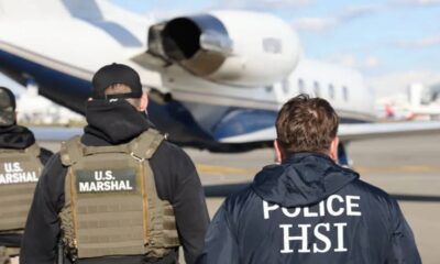 U.S. Marshals and HSI officers walk toward a private jet on the airport tarmac, seen from the back with tactical gear on.