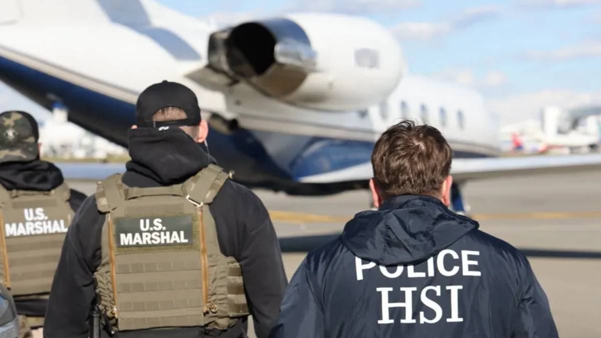 U.S. Marshals and HSI officers walk toward a private jet on the airport tarmac, seen from the back with tactical gear on.