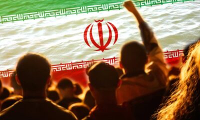 Silhouetted crowd at an outdoor event with a large Iranian flag overhead, green-white-red with the emblem visible in the center.