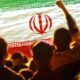 Silhouetted crowd at an outdoor event with a large Iranian flag overhead, green-white-red with the emblem visible in the center.