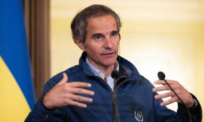 Man in a blue jacket speaks at a press briefing, gesturing with his hands as two microphones are near him; Ukrainian flag in the background.