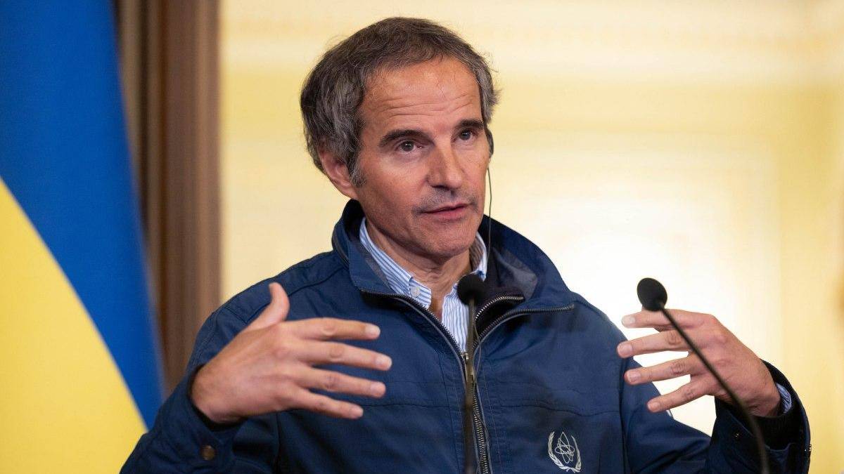 Man in a blue jacket speaks at a press briefing, gesturing with his hands as two microphones are near him; Ukrainian flag in the background.