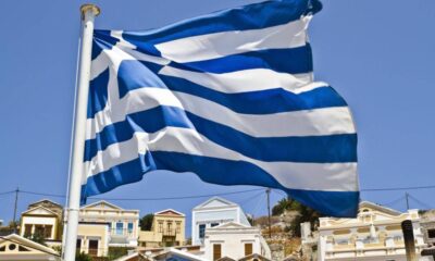 Waving Greek flag on a flagpole with white and blue stripes over a sunny Greek village backdrop