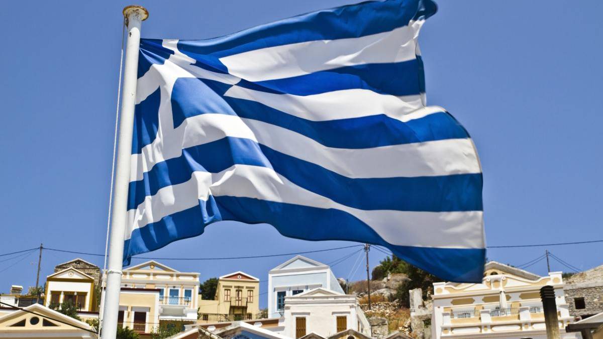 Waving Greek flag on a flagpole with white and blue stripes over a sunny Greek village backdrop