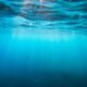 Underwater view looking up at the sunlit blue surface, with light beams piercing the water and small bubbles or particles in the water.