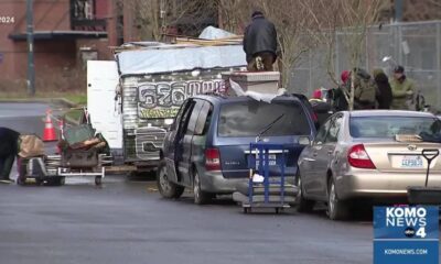 Man standing on top of a graffiti-covered trailer loaded with debris beside a blue minivan and a silver sedan on a city street; several people and a cart nearby (KOMO News).